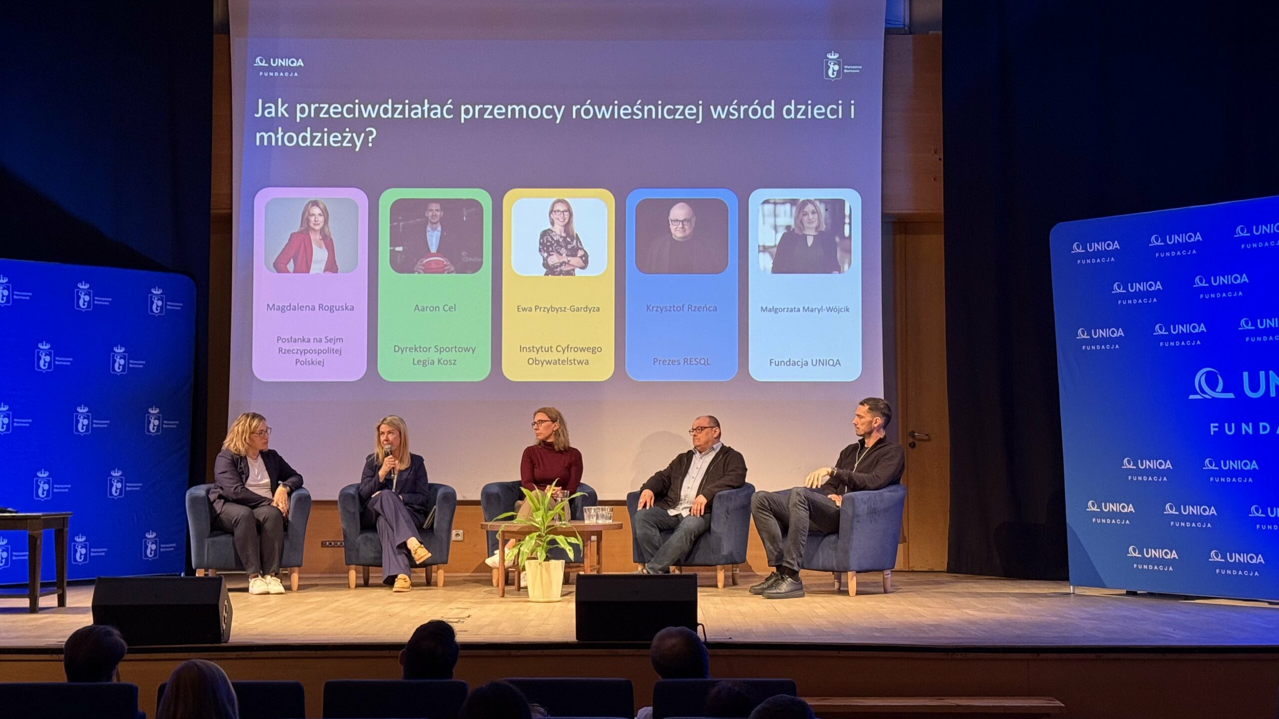 Sala konferencyjna, na której siedzą ludzie na krzesłach, za nimi prezentacja